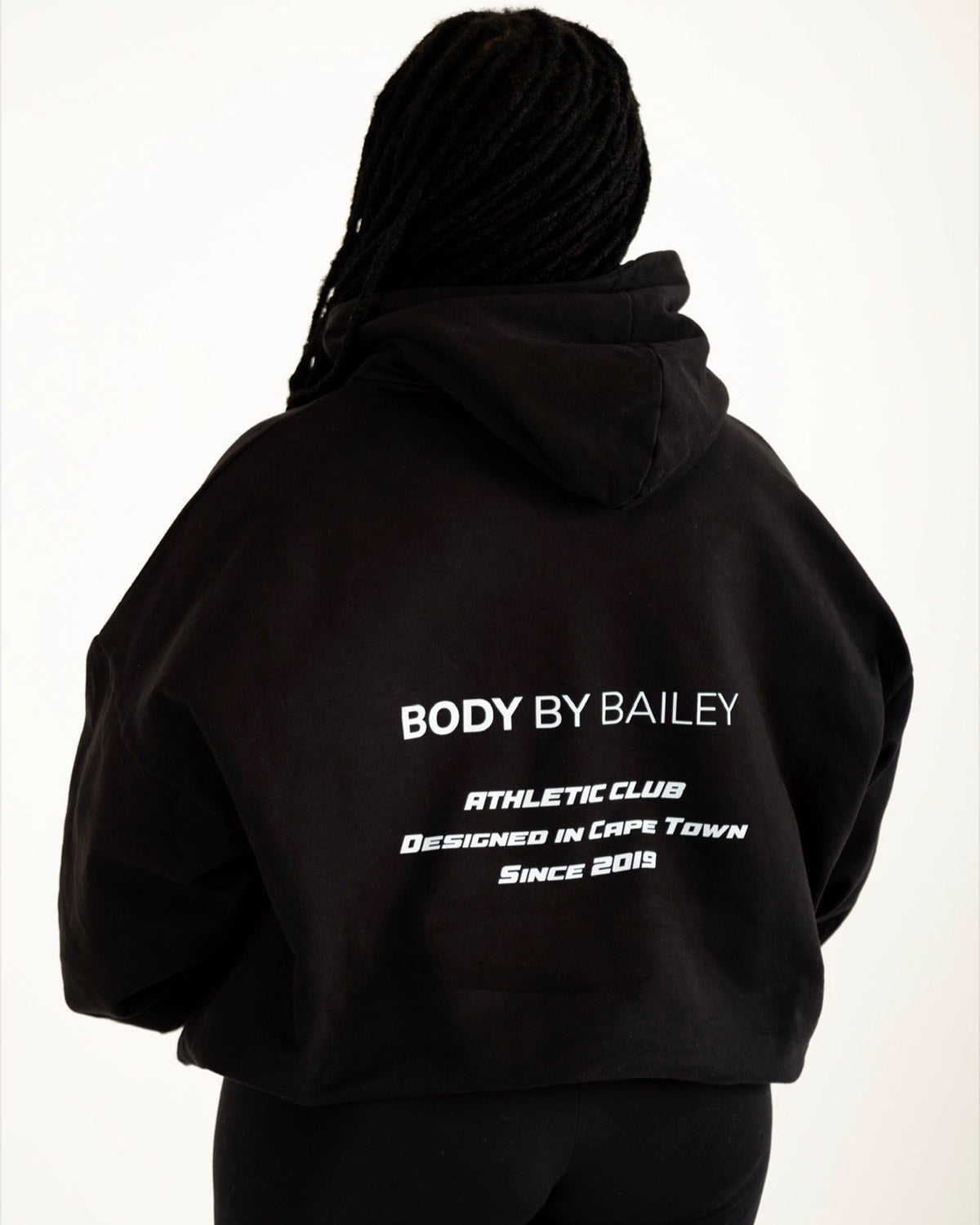 A person shown from the back wearing a black cropped hoodie with white text reading 'BODY BY BAILEY ATHLETIC CLUB DESIGNED IN CAPE TOWN Since 2018' and black leggings against a plain white background.