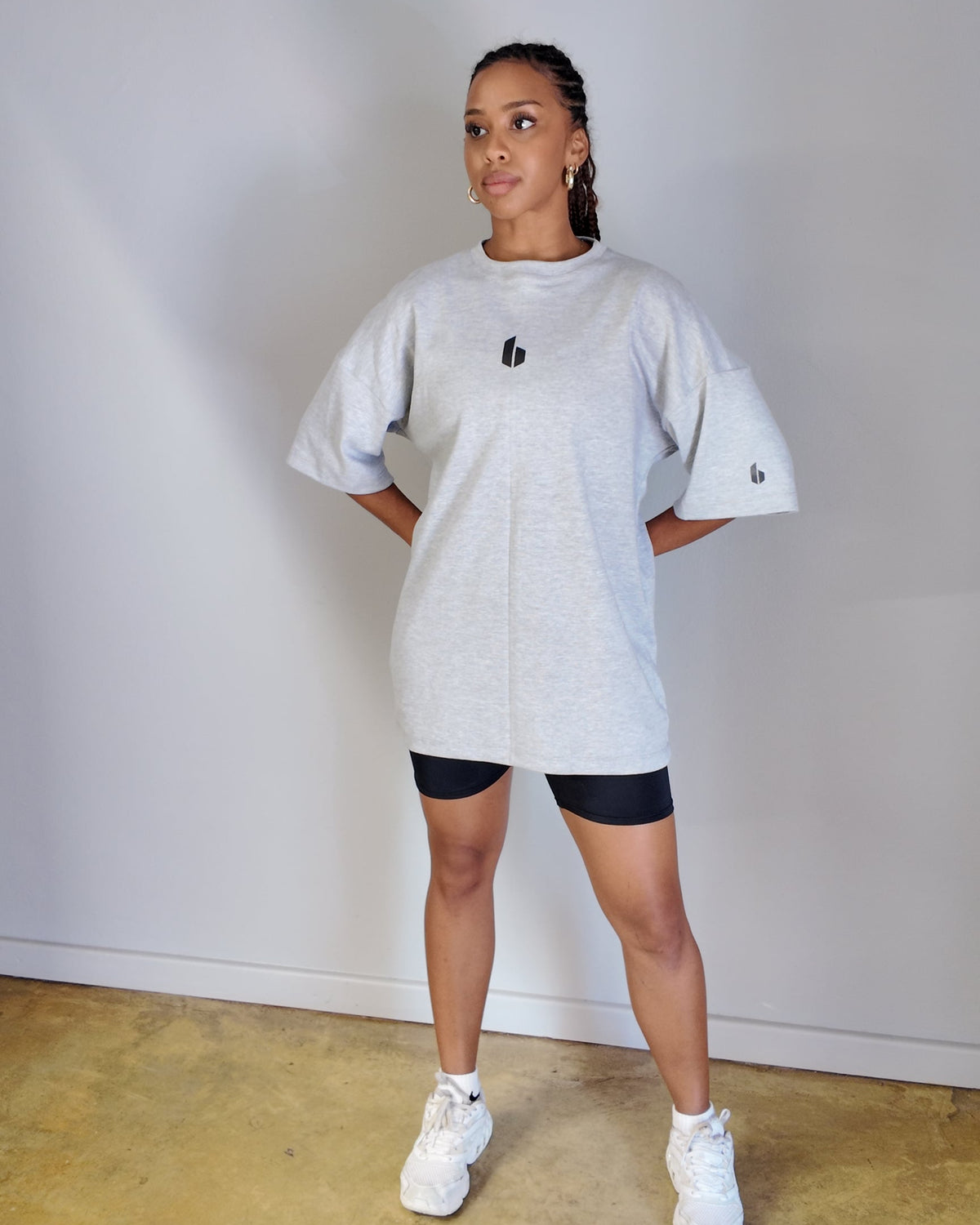 A person wearing a round neckline, oversized, grey melange t-shirt with a small logo on the chest, paired with black shorts and white sneakers.
