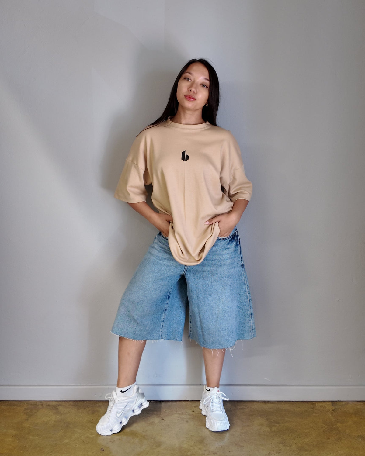 A person wearing an oversized nude-colored t-shirt with a round neckline and a small logo on the chest, paired with blue denim shorts and white sneakers.