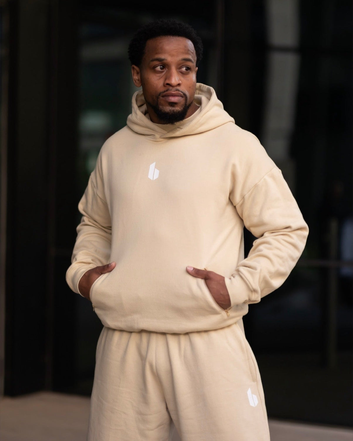 A man wearing a matching beige fleece hoodie and sweatpants set with a small white logo on the chest and thigh, standing with hands in his pockets against a dark modern building background.