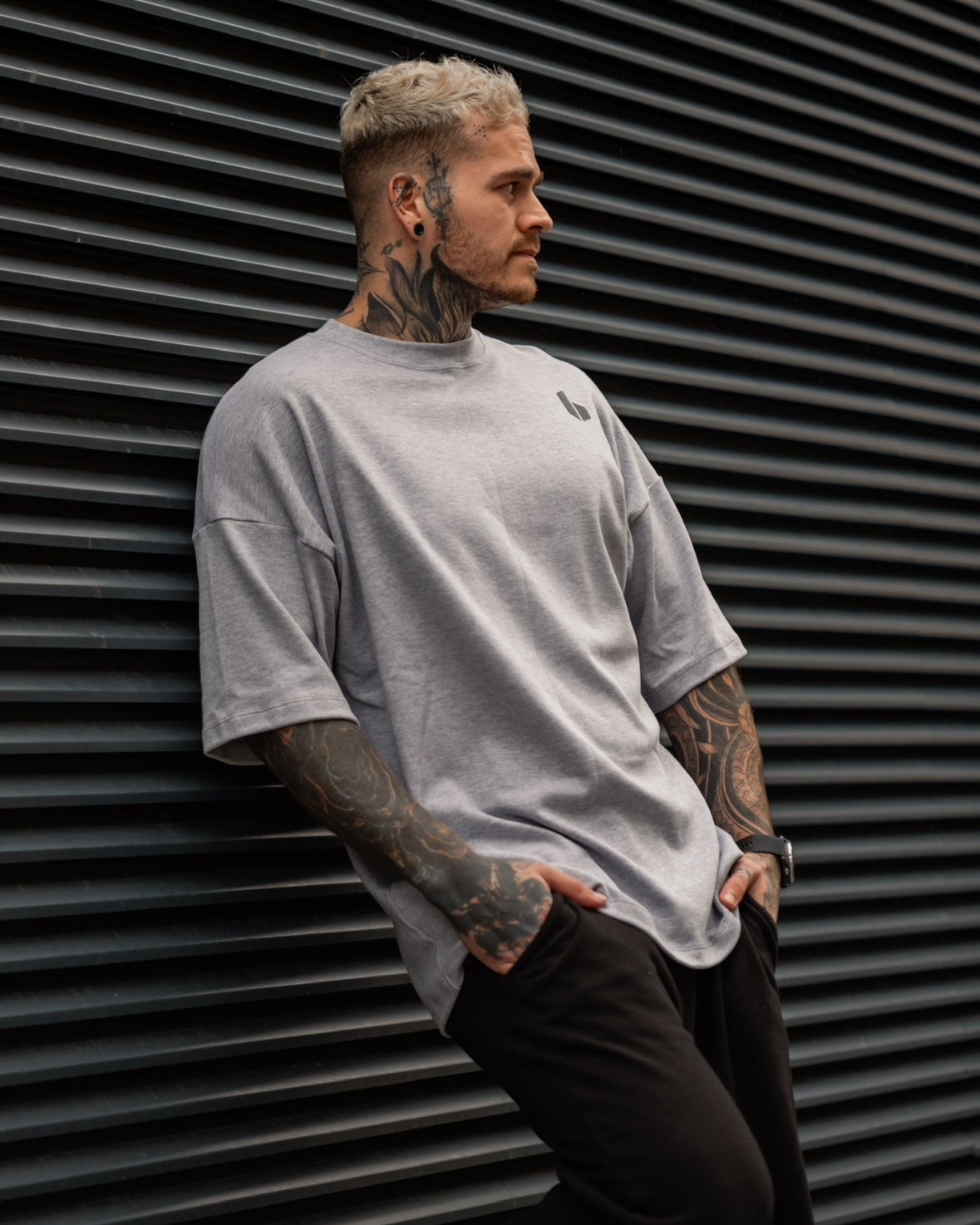 A person with tattoos wearing a grey, oversized round-neck t-shirt with a small logo on the chest, leaning against a dark wall.
