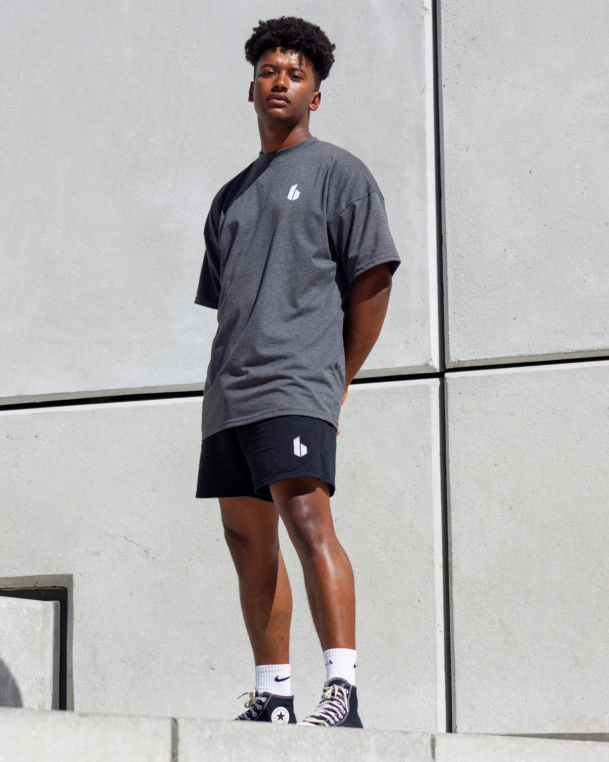 A man standing with one hand on a wall, wearing a charcoal grey oversized T-shirt and black shorts.