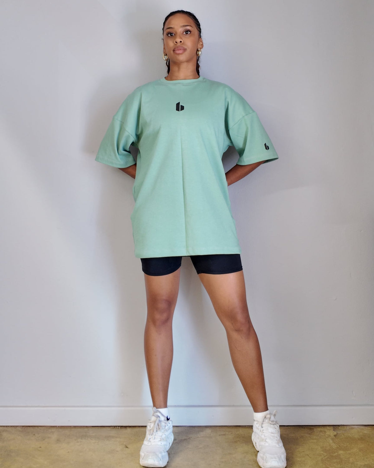 A person wearing an oversized green t-shirt with a round neckline and a small logo on the chest area, paired with black shorts and white sneakers.