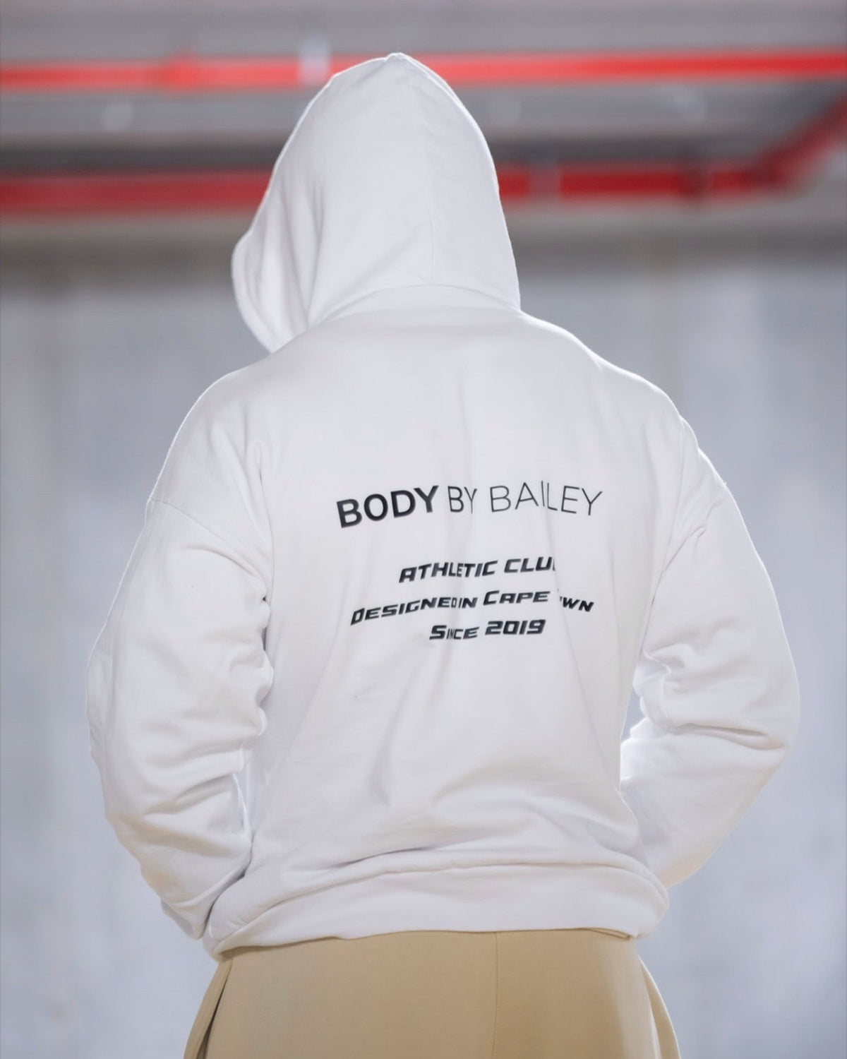 A person viewed from behind wearing a white hooded sweatshirt with black printed text on the back reading 'BODY BY BAILEY' and additional smaller text below, paired with light beige bottoms in an indoor setting with a blurred gray background.