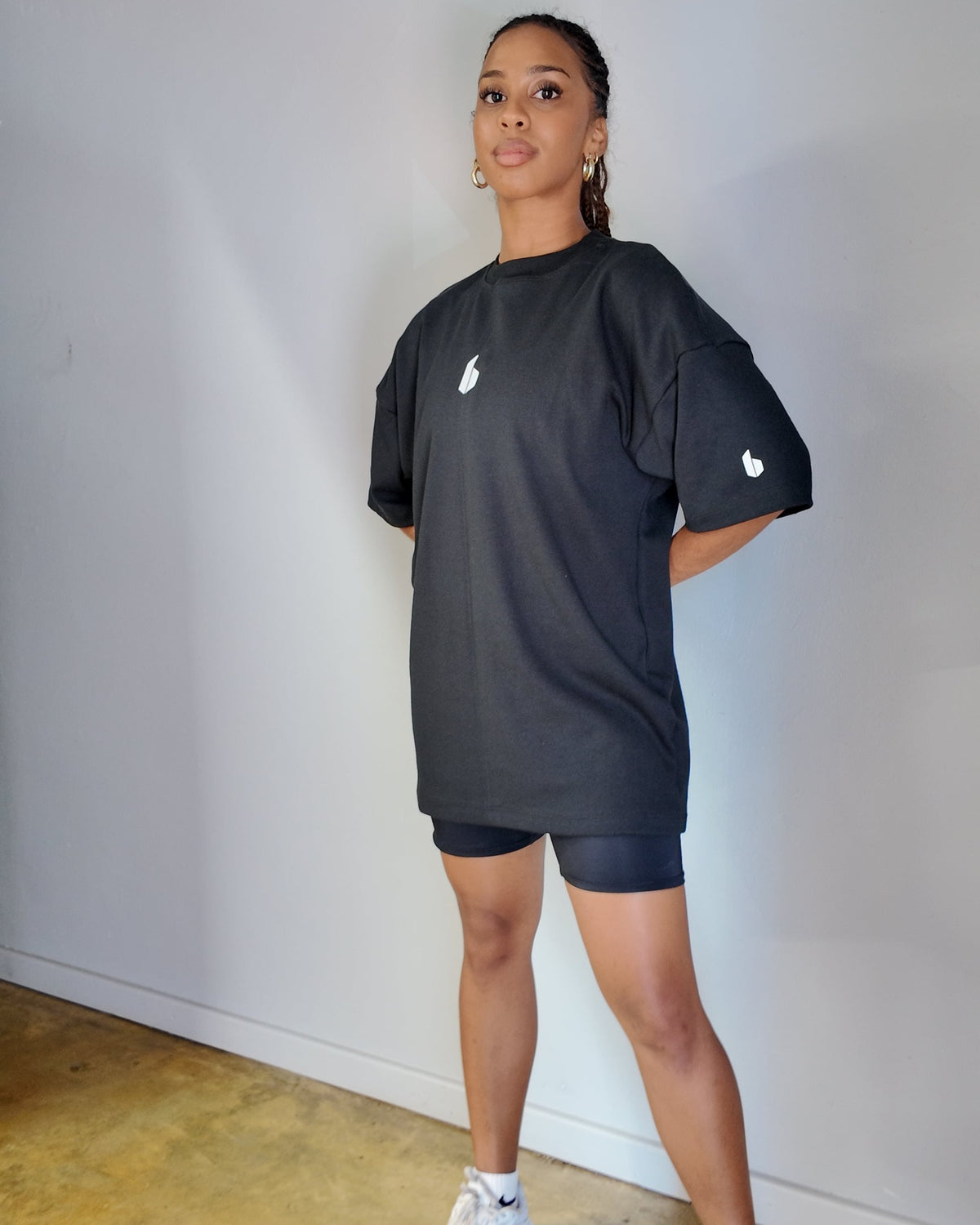 A person wearing a round neckline, oversized black tee with a logo detail on the chest and shorts, standing against a neutral background.