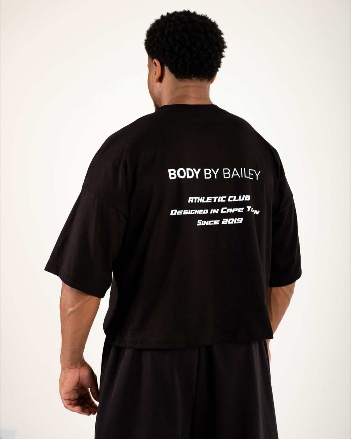 A man shown from behind wearing a black t-shirt and black shorts against a plain light background. The back of the t-shirt features white text that reads 'BODY BY BAILEY' with smaller text below.