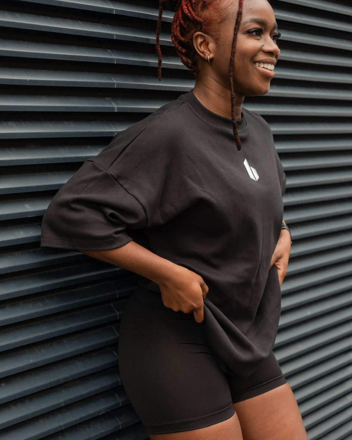 Person wearing a black outfit with a logo against a corrugated metal background
