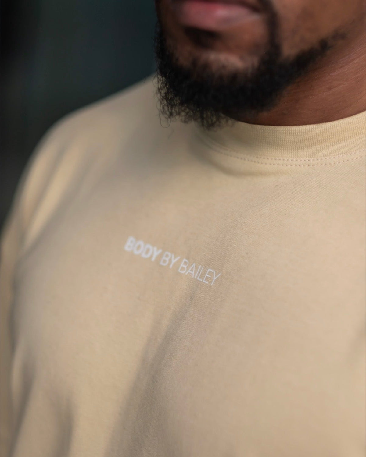 Close-up of a man wearing a beige crewneck sweatshirt with 'BODY BY BAILEY' printed in white on the chest.