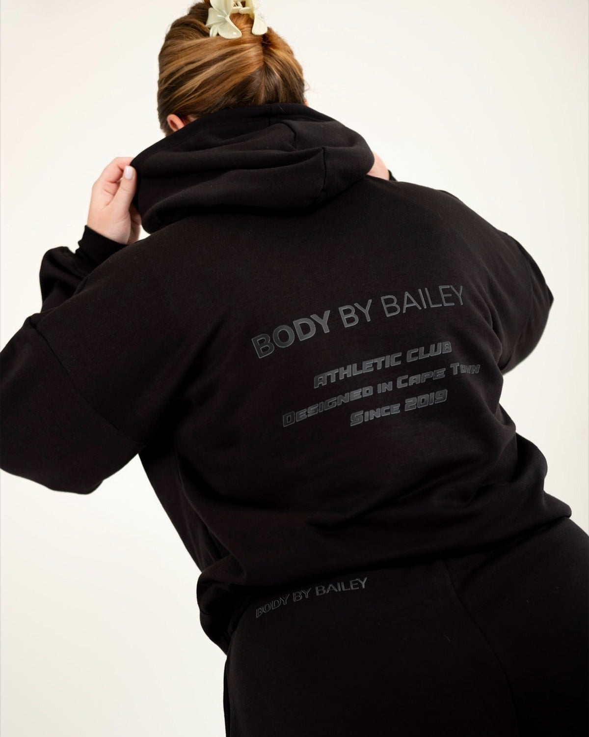 A person wearing a black hoodie and matching black sweatpants, photographed from the back against a plain light background. The hoodie has gray text on the back reading 'BODY BY BAILEY' with smaller text below.