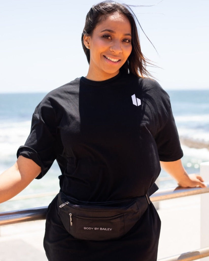 A person standing by the sea wearing a black oversized crew neck t-shirt with short sleeves and a front pocket, with the brand logo visible on the shirt.