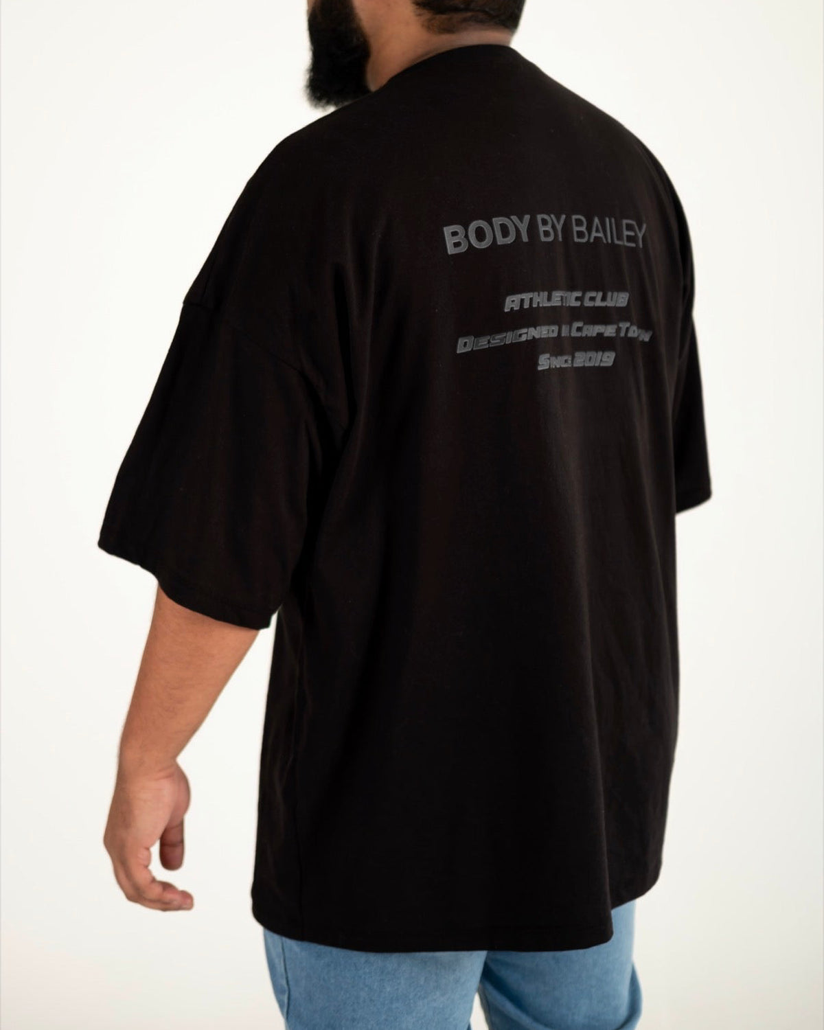 A man wearing an oversized black t-shirt from the back, featuring white text that reads 'BODY BY BAILEY ATHLETIC CLUB' and 'GOLDEN WOOD CAFE & TEE SHOP'. He is also wearing a white cap and light blue jeans, photographed against a plain white studio background.