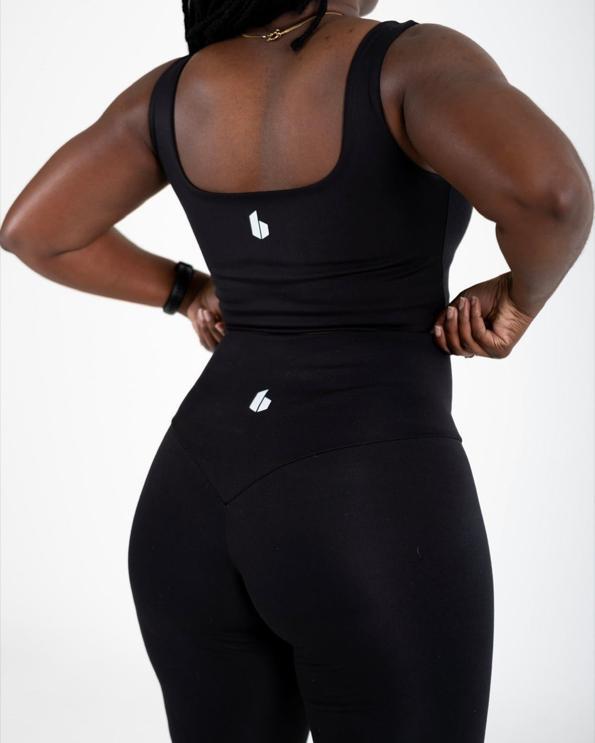 A woman shown from the back wearing a black athletic tank top and matching high-waisted leggings with small white logo details, against a plain light gray studio background.
