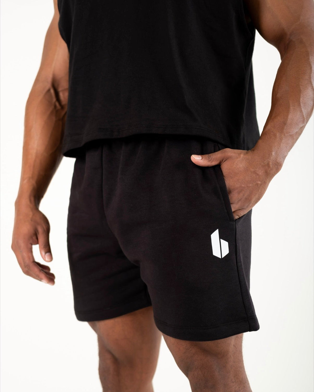 A close-up of a person wearing black athletic shorts with a white geometric logo on the left leg, paired with a black sleeveless top against a plain light background.