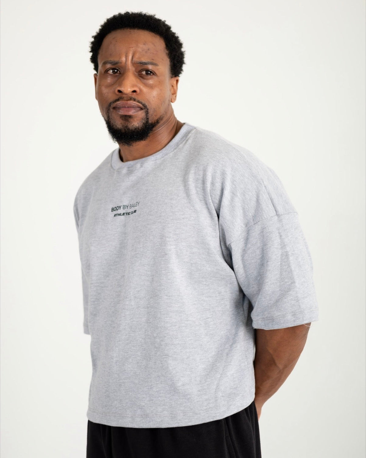 A man wearing a light gray oversized crewneck t-shirt with small black text on the left chest, paired with black shorts, photographed against a plain light gray studio background.