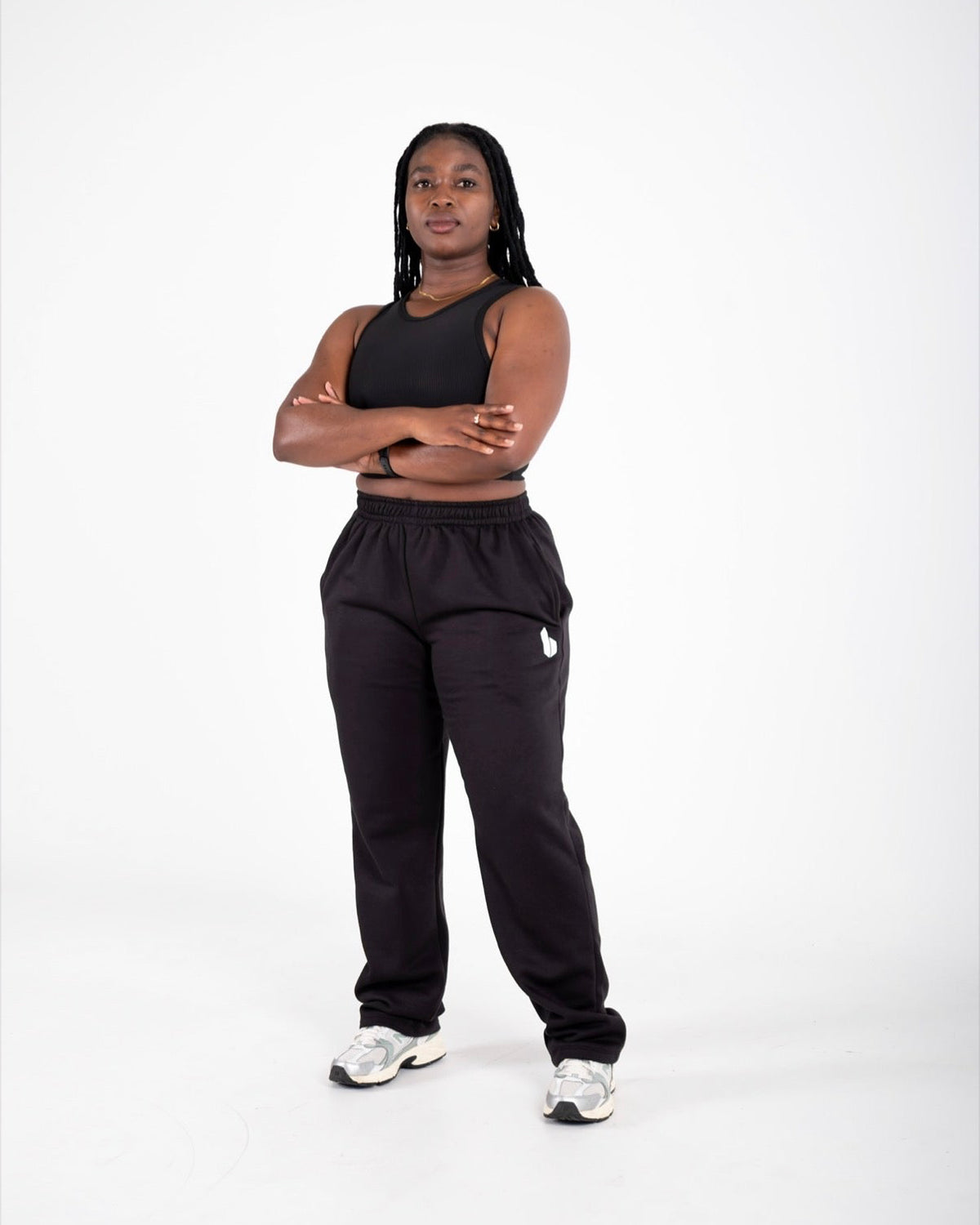 A woman modeling a black athletic set consisting of a sleeveless crop top and matching sweatpants, standing against a seamless white studio background.