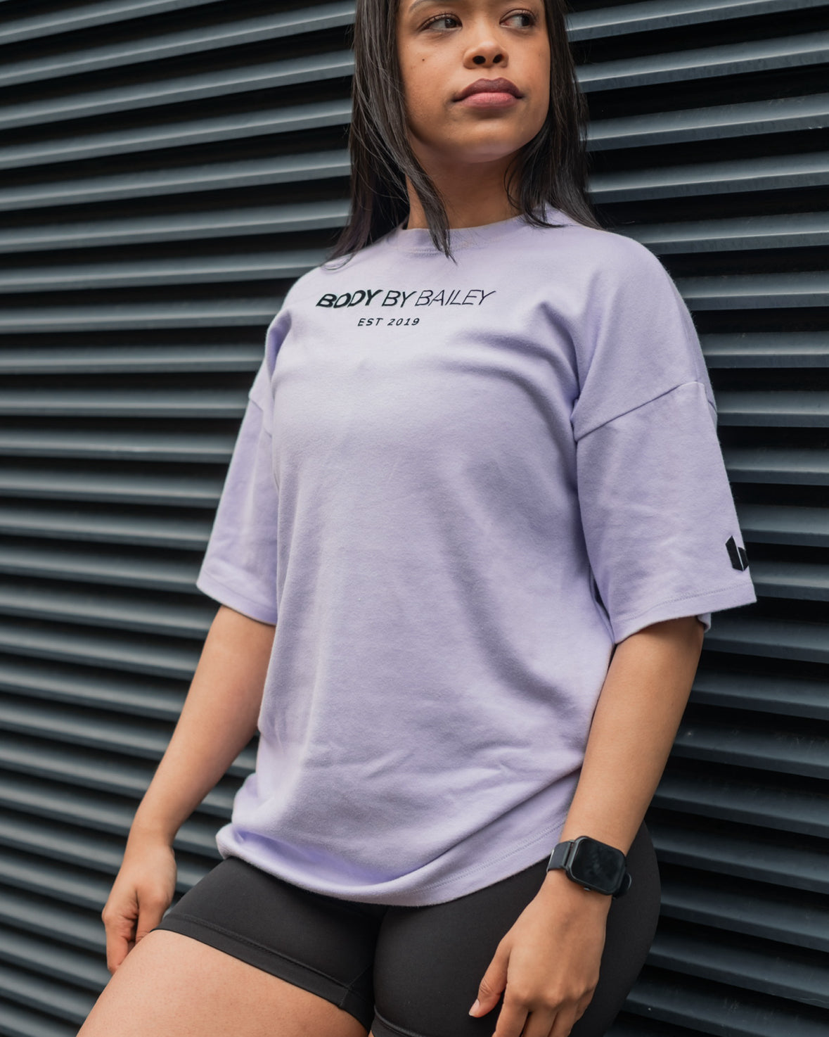 A person wearing an oversized lilac t-shirt with a round neckline and short sleeves, standing in front of a corrugated metal background.