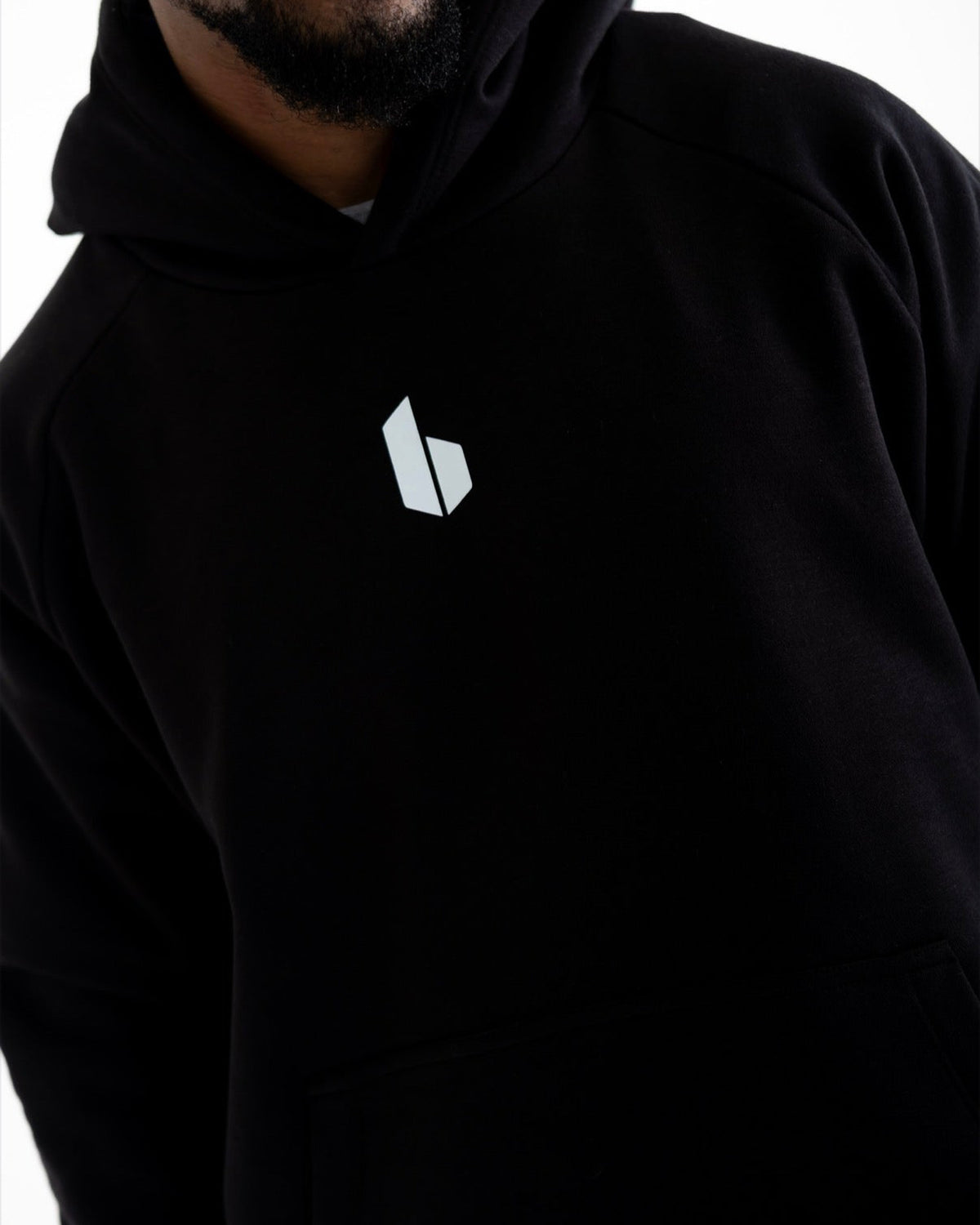 A close-up of a man wearing a black pullover hoodie with a small white geometric logo on the left chest, set against a plain white background.