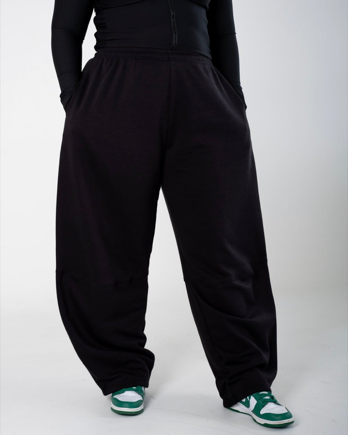 A person wearing loose black sweatpants with a black long-sleeve top and green and white sneakers, shown from the chest down against a light gray studio background.