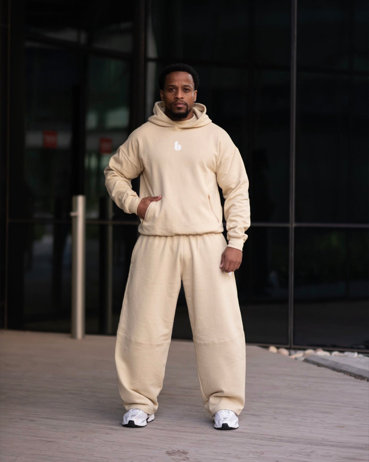 A man wearing a matching beige hoodie and sweatpants set with white sneakers, standing in front of a modern glass building.