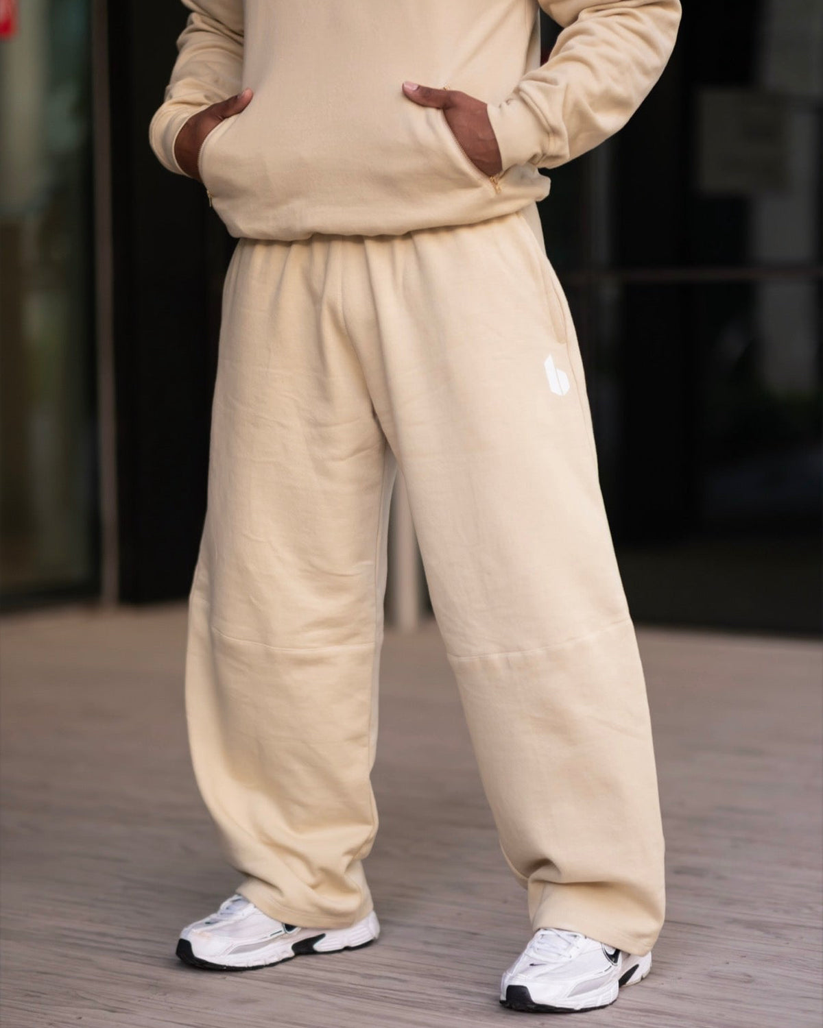 A person wearing a matching beige sweatshirt and loose-fit jogger pants, standing on a gray paved surface with hands in the sweatshirt pockets. The pants have a small white graphic on the upper left thigh.