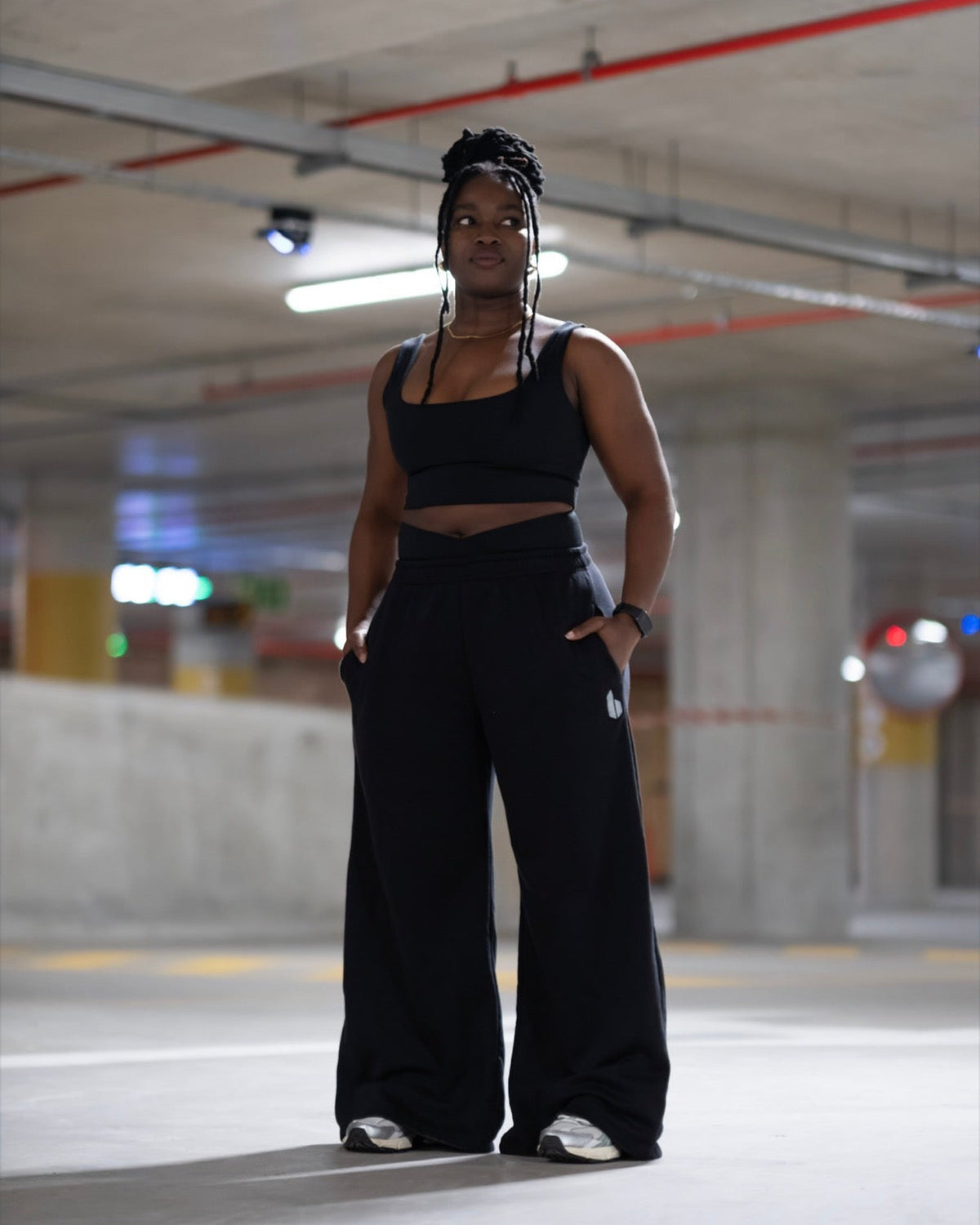 A woman stands in a concrete parking garage wearing a black two-piece activewear set consisting of a cropped tank top and matching wide-leg sweatpants.